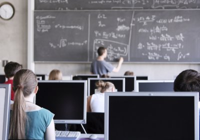 Auf dem Bild zu erkennen sind Schülerinnen und Schüler in einer Unterrichtsstunden am PC. Ein Lehrer schreibt Formeln an die Tafel.
