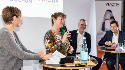 Staatssekretärin Andrea Milz mit einem Mikrofon in der Hand sitzt mit Teilnehmern der Pressekonferenz auf Hockern an Rundtischen. Hinter ihnen eine weiße Rückwand mit einer Präsentation.
