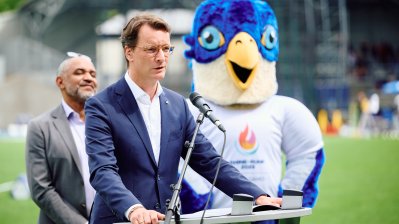 MP Wüst hält ein Grußwort, im Hintergrund das Maskottchen