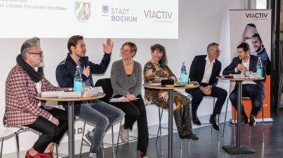 Staatssekretärin Andrea Milz sitzt mit Teilnehmern der Pressekonferenz auf Hockern an Rundtischen. Hinter ihnen eine weiße Rückwand mit einer Präsentation.