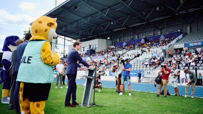 MP Wüst hält ein Grußwort, im Vordergrund das Maskottchen