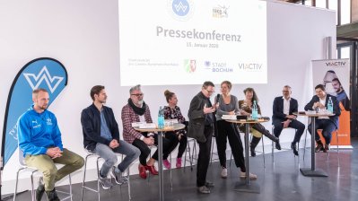 Staatssekretärin Andrea Milz sitzt mit Teilnehmern der Pressekonferenz auf Hockern an Rundtischen. Hinter ihnen eine weiße Rückwand mit einer Präsentation.