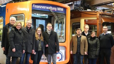 Ein Gruppe Menschen ssteht vor einer U-Bahn, die mit der Werbung der Finanzverwaltung NRW verziert ist