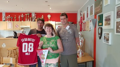 Staatssekretärin Andrea Milz mit zwei Vereinsmitgliedern im Clubraum, neben sich ein roten Vereinstrikot. Hinten an der Rückwand stehen viele Pokale und rechts an der Wand mehrere eingerahmte Fotos.