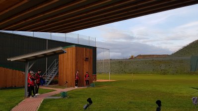 Blick ins Stadion. Grüner Rasen, Tribünen, links ein Holzgebäude, oben eine Überdachung.