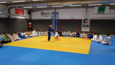 Zwei jugendliche Judokas demonstrieren Selbstverteidigung auf einer gelben, blau-umrandeten Matte. Auf dem breiten blauen Rand sitzen im Halbkreis  weitere Judokas. Rechts am Bildrand steht der Trainer.