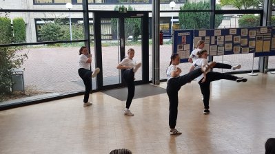 Fünf junge Frauen in schwarzen Jogginghosen und weißen T-Shirt beim Selbstverteidigungstraining, dabei mache sie alle gleichzeitig mit dem rechten Bein einen hohen Seitwärts-Kick.