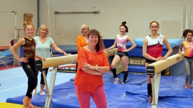 Staatssekretärin Milz in orangefarbenem T-Shirt und Sporthose mit sechs Damen in einer Turnhalle. Zwischen den Damen stehen zwei Schwebebalken, auf dem Boden liegen blaue Bodenmatten.