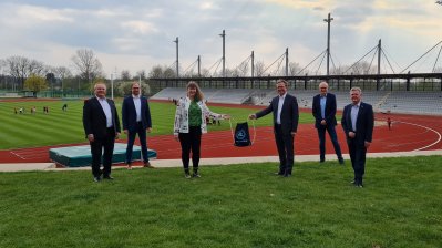 Staatssekretärin Andrea Milz steht mit 5 weiteren Menschen vor einer Sportstätte mit Tartanbahnen und einem Rasenplatz in der Mitte.