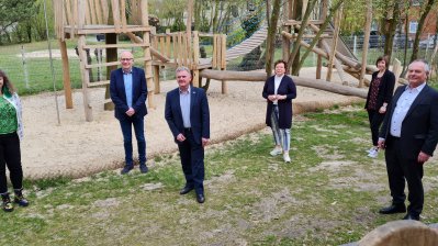 Staatssekretärin Andrea Milz steht mit 5 weiteren Menschen auf einer Wiese vor einem Sandspielplatz mit vielen Holzgerüsten.