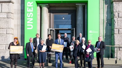 Eine Gruppe Menschen steht vor einem Eingang mit grünem Tor, zwei halten jeweils eine große Karte