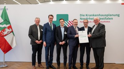 MP Wüst und Minister Lauman posieren mit Förderbescheidempfängern und eine Urkunde für ein Foto