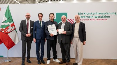 MP Wüst und Minister Lauman posieren mit Förderbescheidempfängern und eine Urkunde für ein Foto