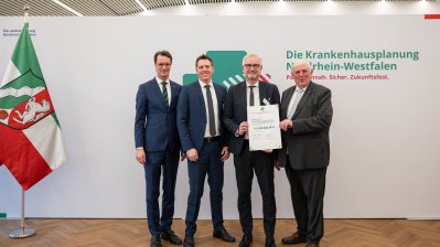 MP Wüst und Minister Lauman posieren mit Förderbescheidempfängern und eine Urkunde für ein Foto
