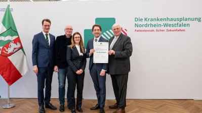 MP Wüst und Minister Lauman posieren mit Förderbescheidempfängern und eine Urkunde für ein Foto