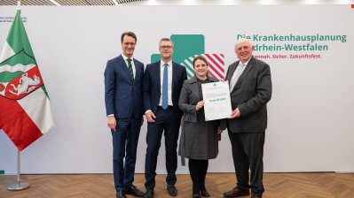 MP Wüst und Minister Lauman posieren mit Förderbescheidempfängern und eine Urkunde für ein Foto