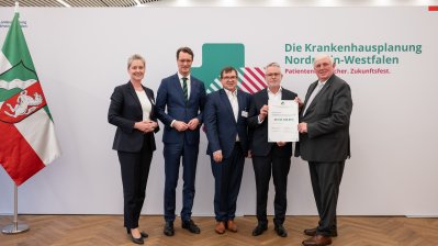 MP Wüst und Minister Lauman posieren mit Förderbescheidempfängern und eine Urkunde für ein Foto