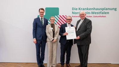 MP Wüst und Minister Lauman posieren mit Förderbescheidempfängern und eine Urkunde für ein Foto