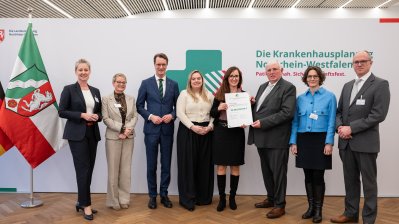 MP Wüst und Minister Lauman posieren mit Förderbescheidempfängern und eine Urkunde für ein Foto