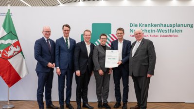 MP Wüst und Minister Lauman posieren mit Förderbescheidempfängern und eine Urkunde für ein Foto