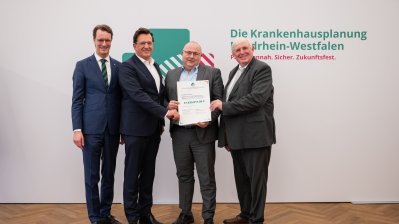 MP Wüst und Minister Lauman posieren mit Förderbescheidempfängern und eine Urkunde für ein Foto