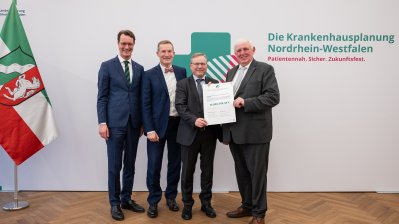 MP Wüst und Minister Lauman posieren mit Förderbescheidempfängern und eine Urkunde für ein Foto