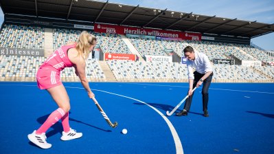 MP Wüst und eine Hockey-Spielerin spielen auf einem blauen Stadionplatz