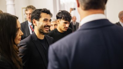 Fußballer İlkay Gündoğan trägt einen dunklen Anzug und lacht zwischen anderen Personen in Business-Kleidung.