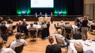 Ein Saal mit vielen, runden, weißen Tischen mit Gästen, weiter vorne ein Bühne  mit einer großen Leinwand.