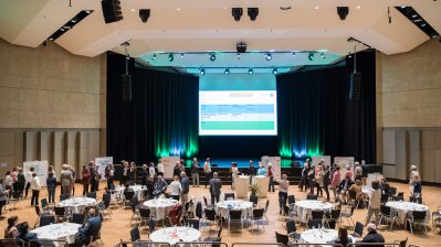 Ein Saal mit vielen, runden, weißen Tischen mit Gästen, weiter vorne ein Bühne  mit einer großen Leinwand.