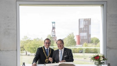 Ministerpräsident und der Oberbürgermeister stehen nebeneinander vor dem Stahlbuch und schauen in die Kamera. Im Hintergrund ist die Zeche Zollverein zu sehen.