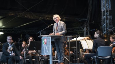 Der Minister steht am Rednerpult, hinter ihm sitzen Musiker des Orchesters