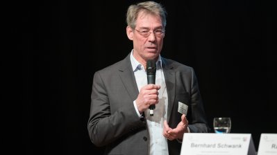 Ein Redner im grauen Anzug und weißem Hemd mit Mikrofon in der Hand am Rednerpult.