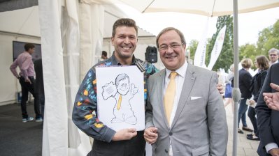 Ein Karikaturist steht mit seiner Zeichnung neben Ministerpräsident Armin Laschet