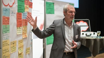 Ein Moderator im grauem Anzug und wießem Hemd zeigt auf eine Pinnwand.