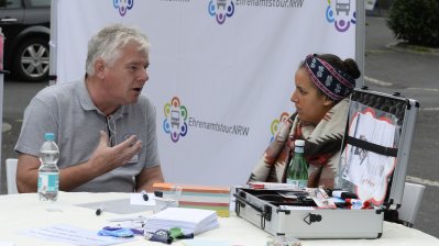 Ein älterer Herr mit einer jungen Dame im Gespräch an einem Tisch sitzend. Im HIntergrund eine Stellwand mit Ehrenamtstour-Logos und dem Ehrenamtstourbus.