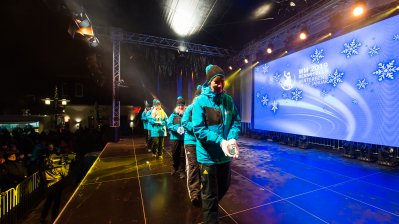 Sporter in türkisfarbenen Jacken gehen mit einem Sportabzeichen vor sich in den Händen quer über die Bühne.