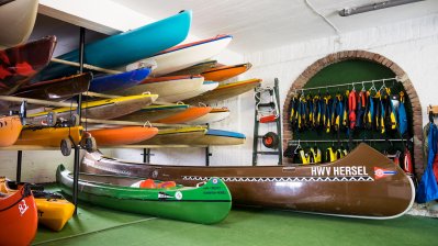 Einblick ins Verinsbootshaus. Verscieden lange Kanus und rechts im Hintergrund verschieden arbige Neoprenanzüge.
