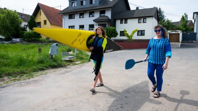 Staatssekretärin Milz mit einem Ruder ihrer rechten Hand geht neben einem jungen Mann, der ein gelbes Kanu trägt, eine Straße entlang.