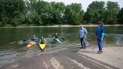 Staatssekretärin Milz steht am Ufer eines Sees. Auf dem See sind drei Kanufahrer. Am gegenüberliegenden Ufer stehen Bäume.