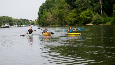 Zwei kanufahrerinnen und ein Kanufahrer rudern über einen Fluss. Im Hintergrund sieht man am linken Ufer ein angelegtes Schiff.