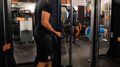 Ein junger Mann in schwarzen T-Shirt und Bermudahose mit weißen Socken und Sportschuhen geht durch einen Fitnessraum.