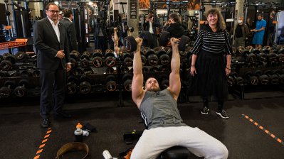 Ein Mann trainiert liegend auf einer Trainingsbank, Staatssekretärin Milz steht rechts daneben.