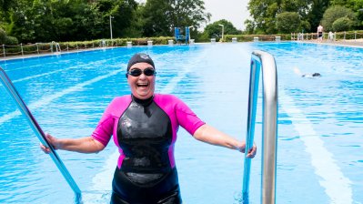 Eine Schwimmerin im pink-schwarzen Neoprenanzug, mit schwarzer Badekappe und schwarzer Schwimmbrille steigt aus einem offenen Schwimmbecken, ihre beiden Hände jeweils links und rechts am Geländer.
