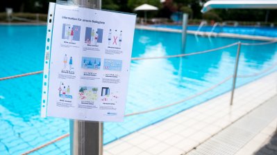 Hygiene-Hinweise in einer Prospekthülle an einem silbernen Mast neben einem offenen Schwimmbecken.