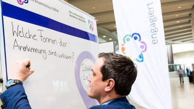 Ein Moderator im blauen Anzug schreibt auf einen Flipchart: "Welche Formen der Anerkennung sind wirksam?"