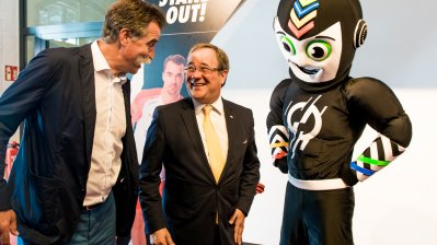 Ministerpräsident Laschet im Gespräch mit einem Mann, rechts neben ihm das Maskottchen.