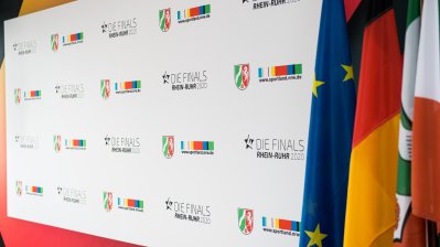 Weiße Plakatwand mit NRW Wappen und Logos von Sportland.NRW, rechts davon hängen die Flaggen von der EU, Deutschalnd und NRW.
