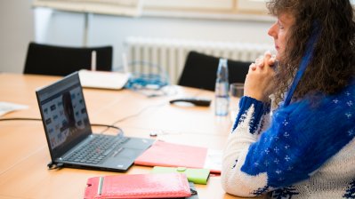 Staatssekretärin Milz im blau-weißen Pullover sitzt an einem beigen Besprechungstisch mit schwarzen Stühlen. Sie schaut vor sich auf einen schwarzen Notebook.