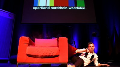 Showakt - Auf der Bühne steht ein großer roter Sessel, rechts daneben liegt ein Mann auf einem Arm abgestüt, den anderen Arm angewinkelt. Er hat ein weißes Oberteil und eine schwarze Hose an.